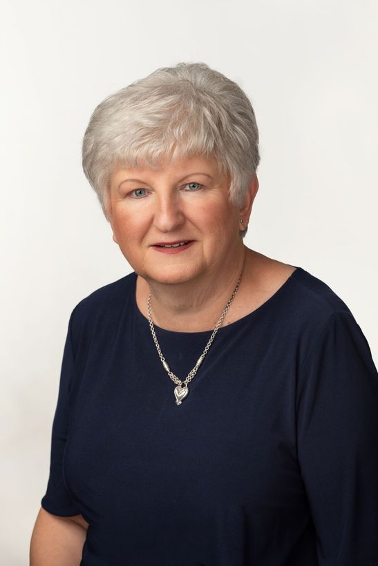 A person with short, grey hair wearing a dark blue top and a silver necklace, smiling against a plain white background.