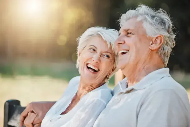 A couple sits on a park bench together, laughing with their heads turned toward each other in a brightly lit setting.