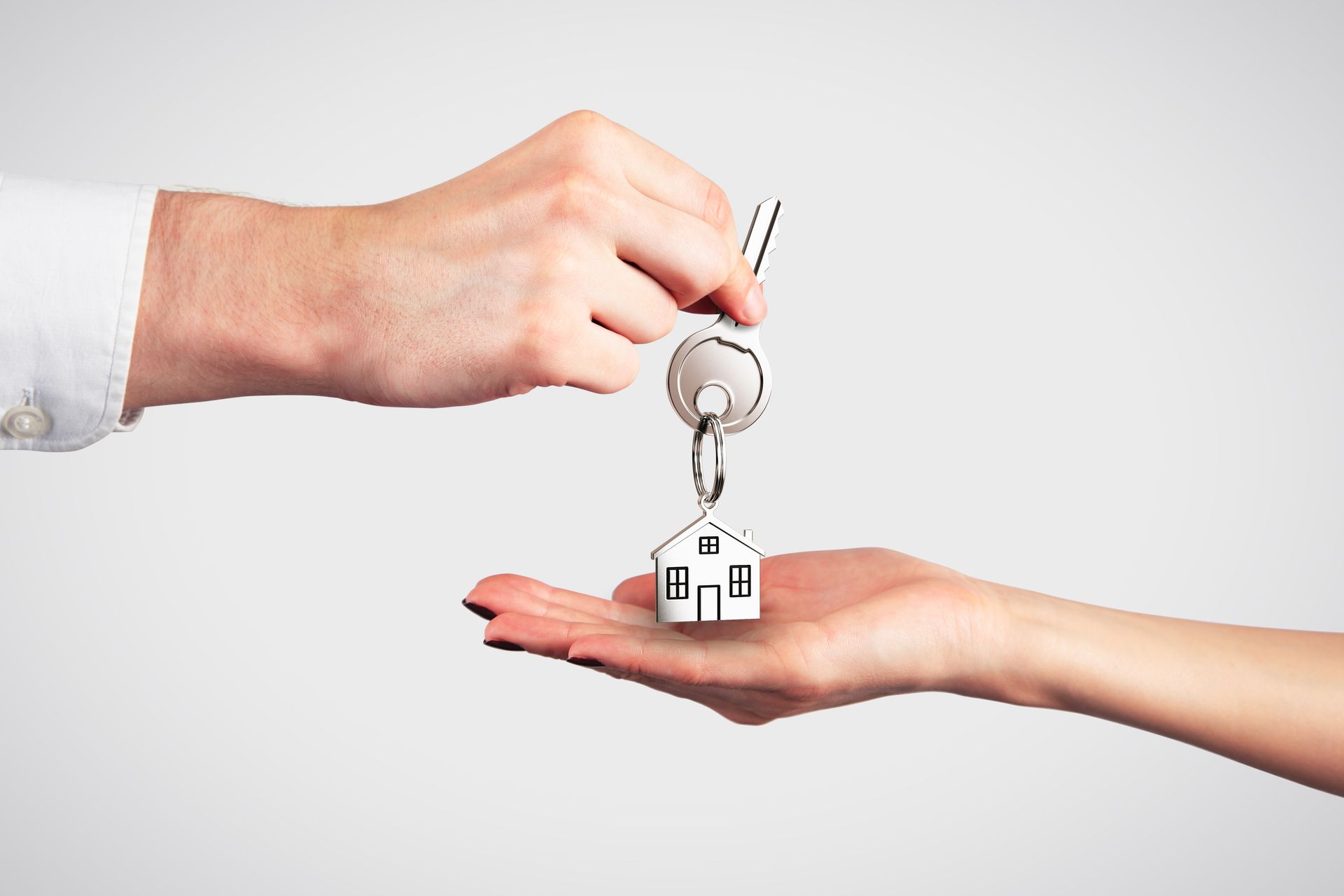A hand hands a key with a small house-shaped keychain to another open, waiting hand against a plain background.