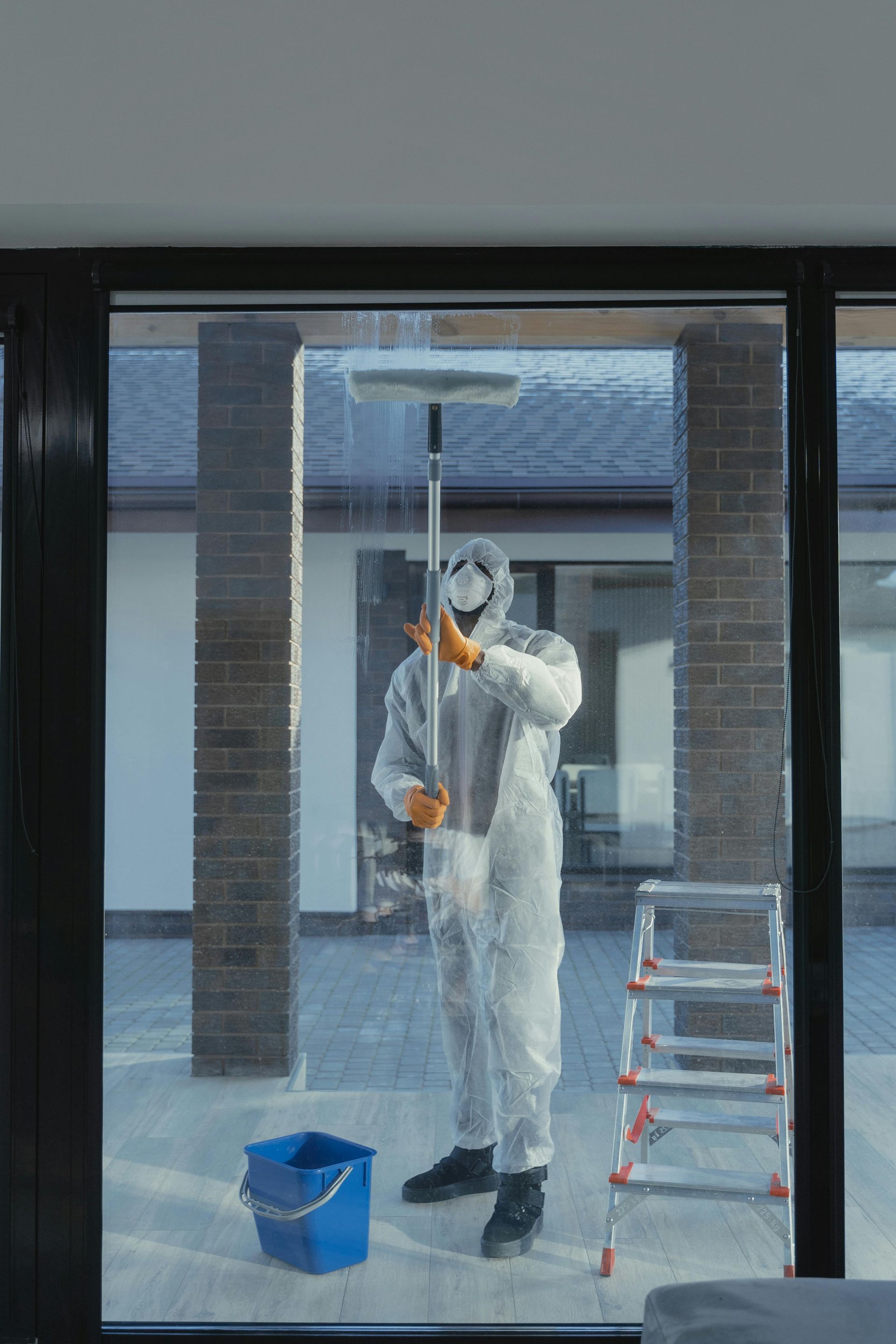 Person in a white hazmat suit cleans a window with a squeegee, standing near a ladder and a bucket outdoors.
