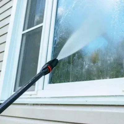 A pressure washer sprays a window, cleaning the glass. The house exterior is visible.