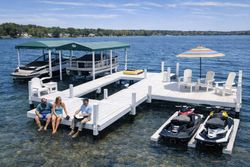 Two people sit on a white dock on a sunny lake, near two covered boats, a shaded seating area, and two jet skis.