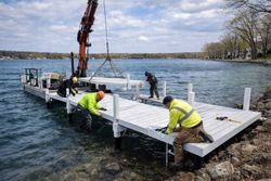 Workers in high-visibility jackets use a barge-mounted crane to install a white dock section in a lake.