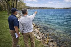 Two people stand on a grassy lakeshore, one pointing toward the water while holding a document.