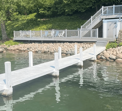 A white wooden dock extending into calm lake water toward a shoreline deck with a stairway and storage shed.