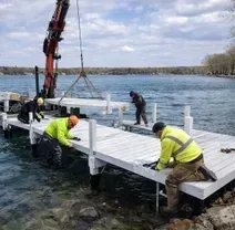 Workers in neon vests use a crane to install a white modular dock into a lake.