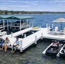 Three people sit on a white dock on a sunny lake, with boats, jet skis, and patio chairs nearby.