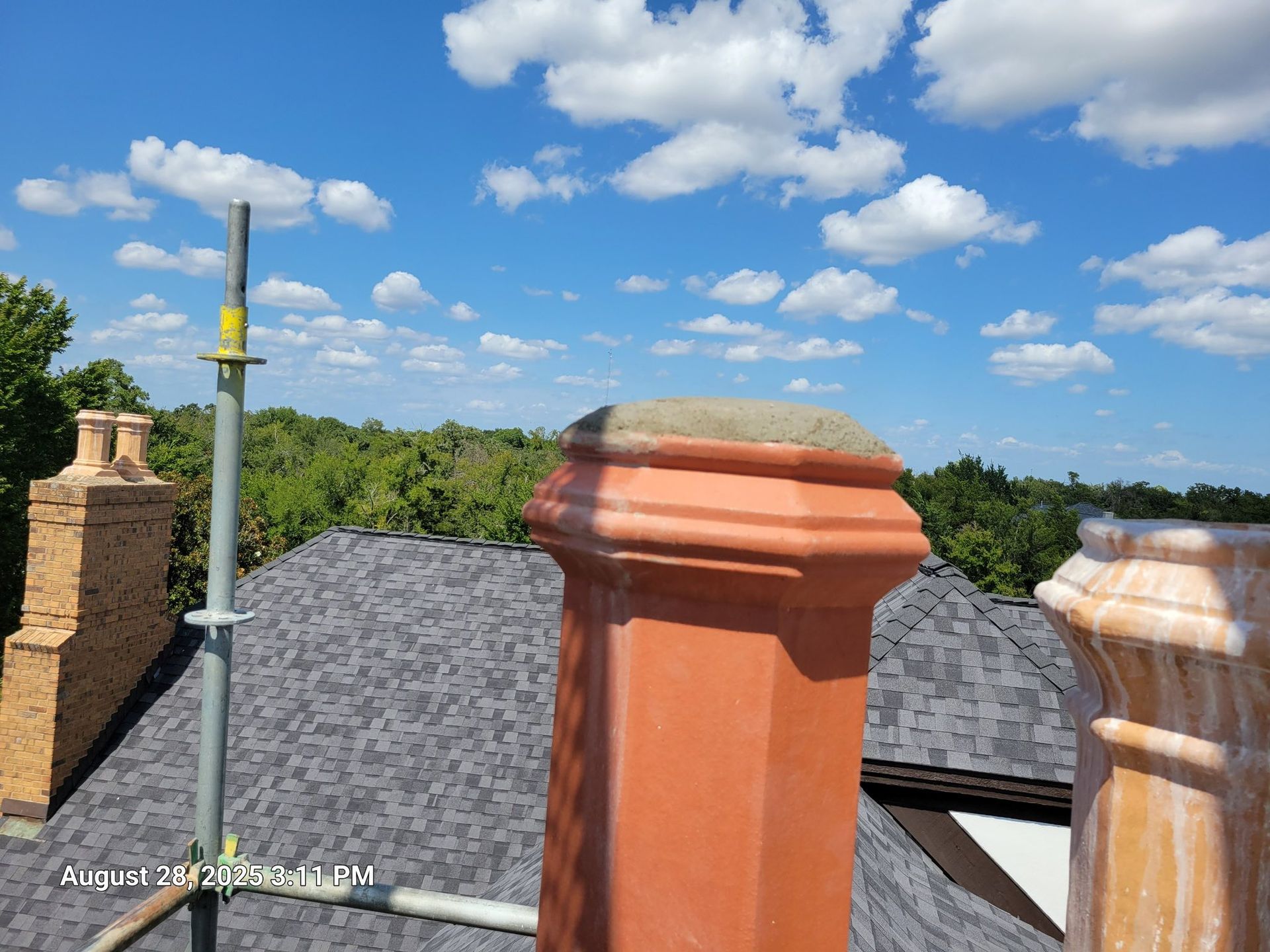 Chimney on a roof against a blue sky with clouds, scaffolding, and trees in the background.