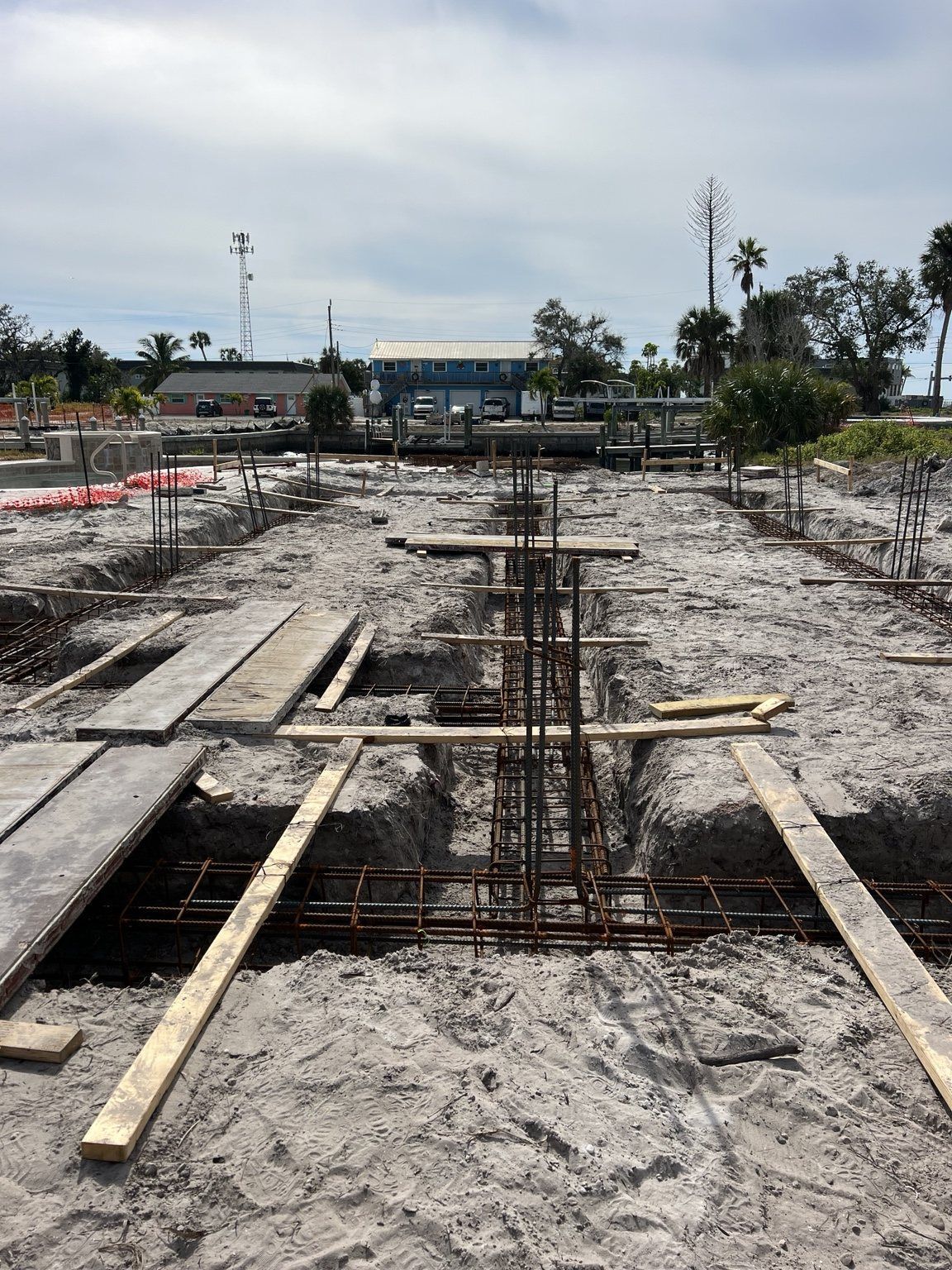 Construction site with rebar foundation footings and wooden forms on sandy ground under a cloudy sky