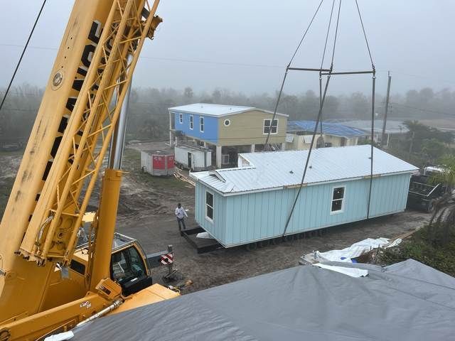 Crane lifting a light blue modular home beside a muddy road in a foggy construction site
