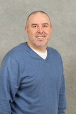 Man with a shaved head and a blue sweater smiles, standing against a gray background.