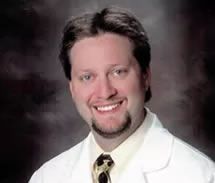 Smiling doctor in white coat, black goatee, yellow tie, against blurred background.
