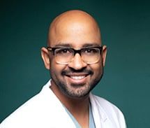 A smiling, bald man wearing glasses and a lab coat against a teal background.