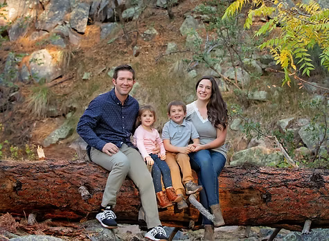 Family of four sitting on a fallen log outdoors in a rocky forest setting.