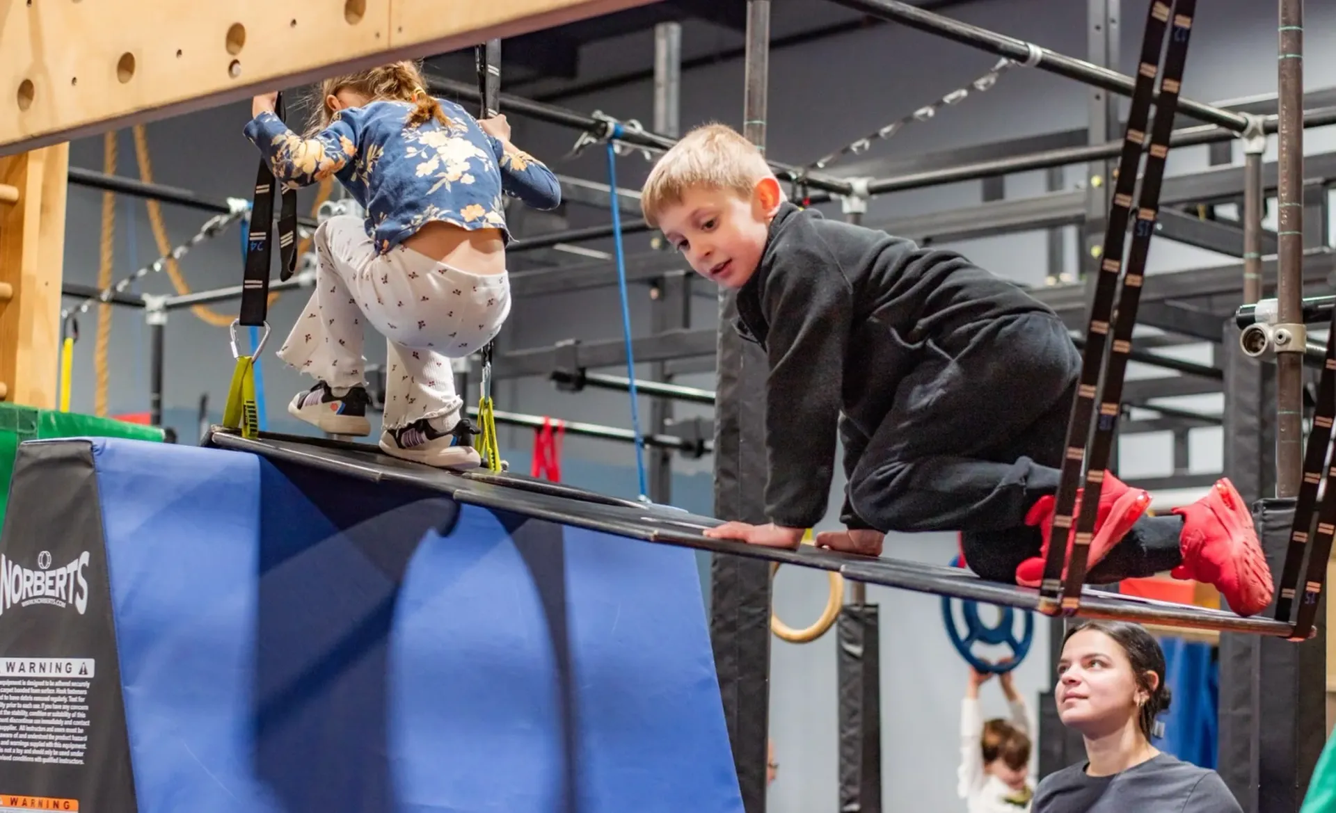 Two children climbing on a black rope obstacle course indoors, with a woman watching nearby.