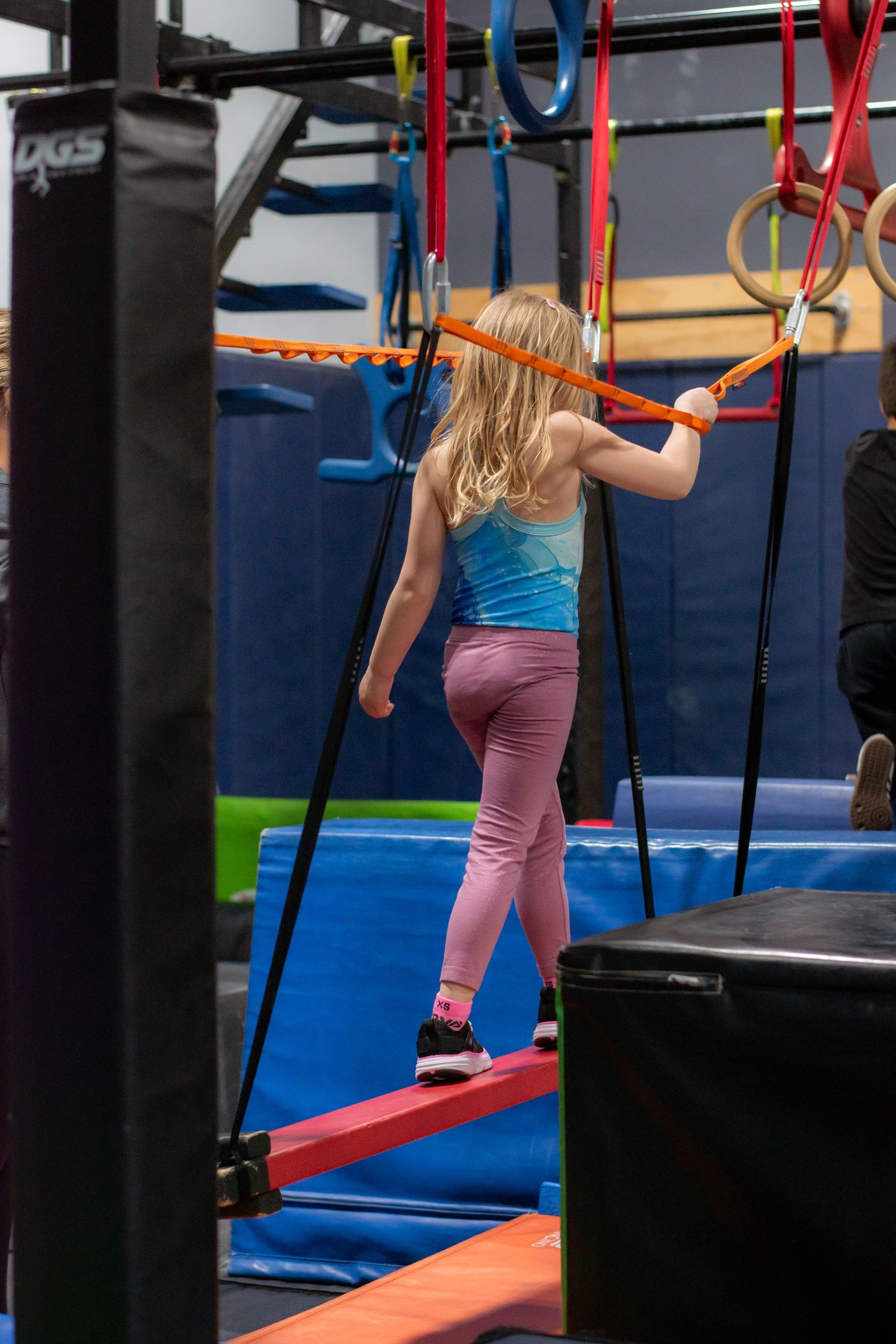 Child balancing on a rope obstacle course in an indoor gym, holding a red rope bridge