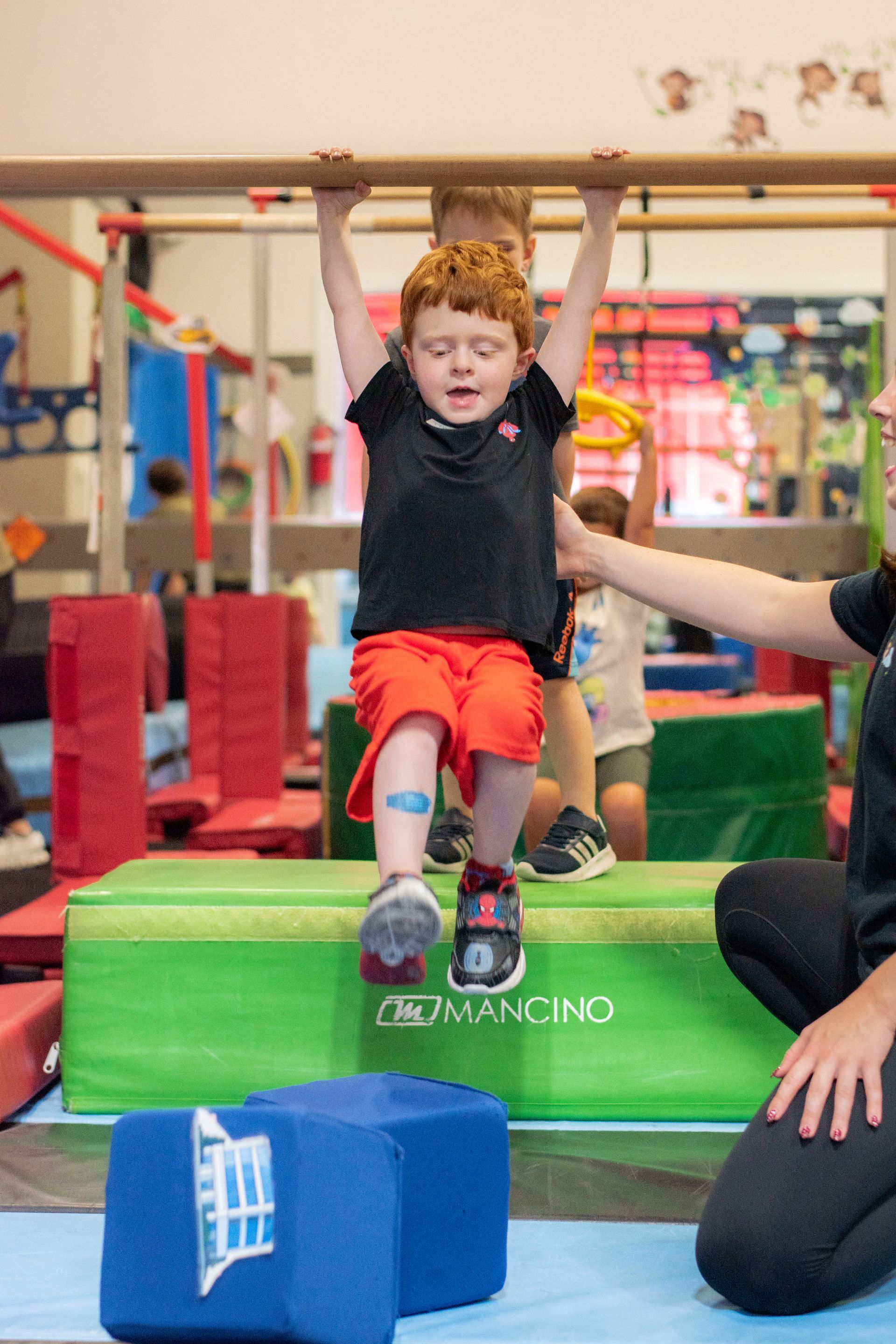 Child swinging on indoor obstacle course with adult nearby, wearing red shorts and black shirt