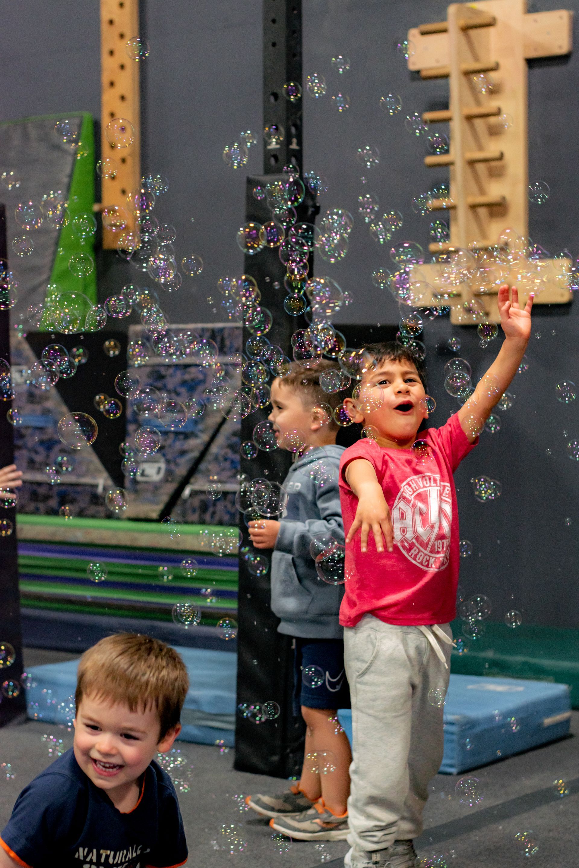 Children playing with bubbles indoors, one reaching up in a pink shirt while others watch nearby