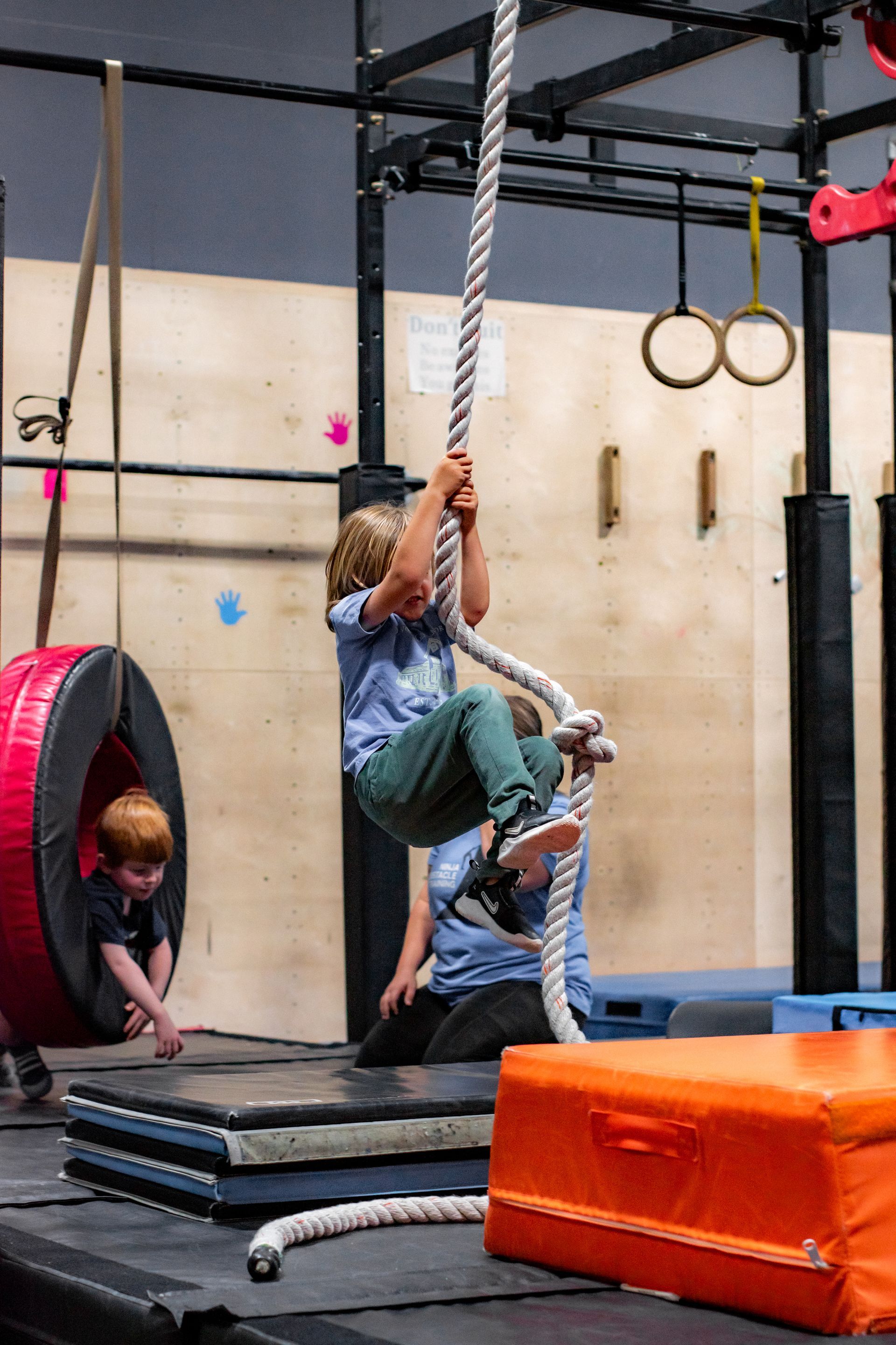 Person climbing a rope in an indoor gym, with mats and gymnastic rings nearby.