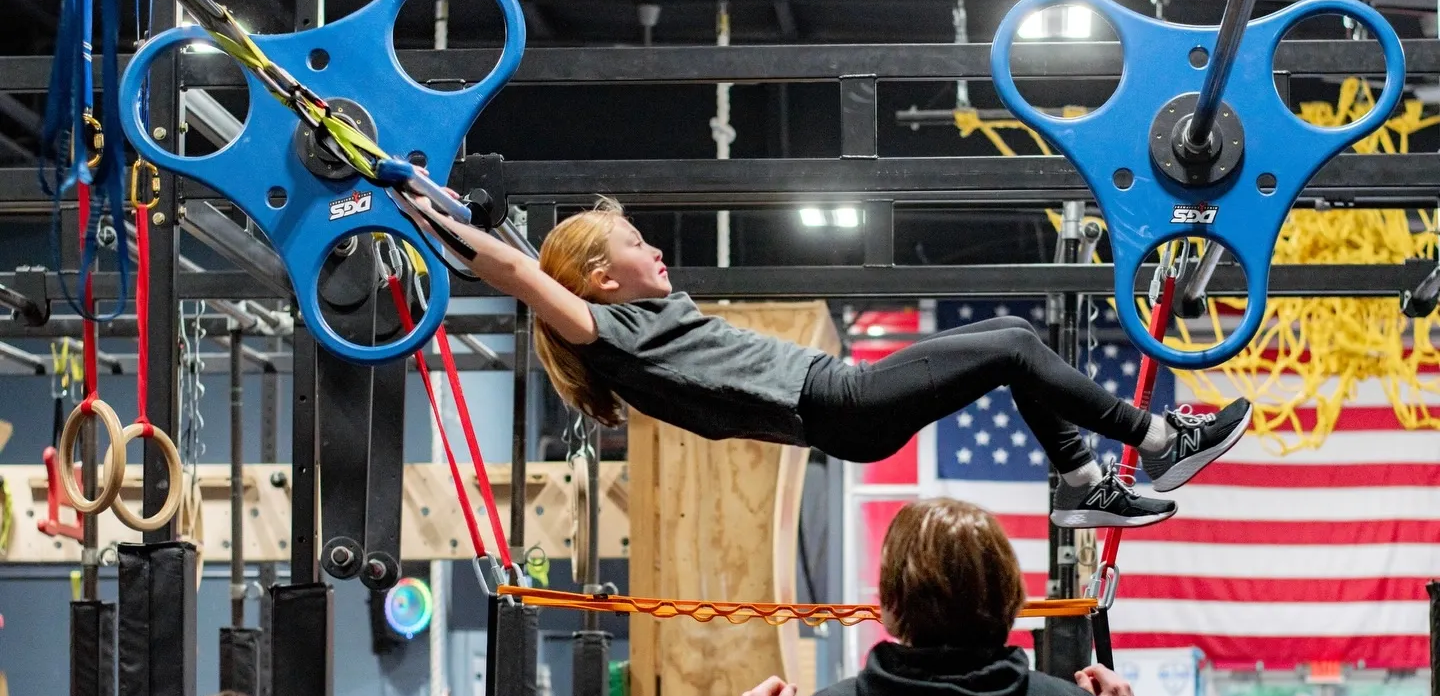 Athlete swinging on blue hanging obstacles in an indoor obstacle course, with an American flag in the background