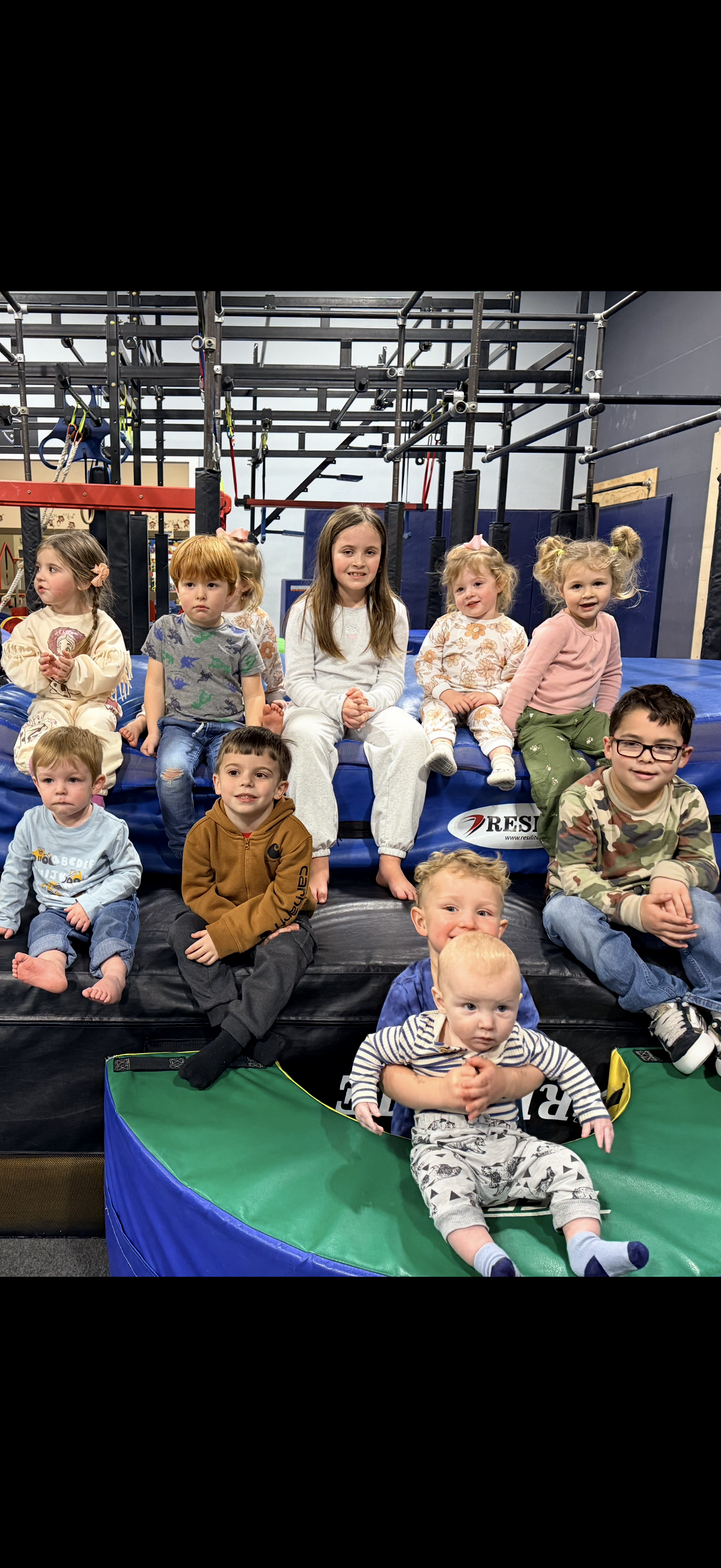 Children sitting on gymnastics mats in a play area, holding small trophies and smiling at the camera