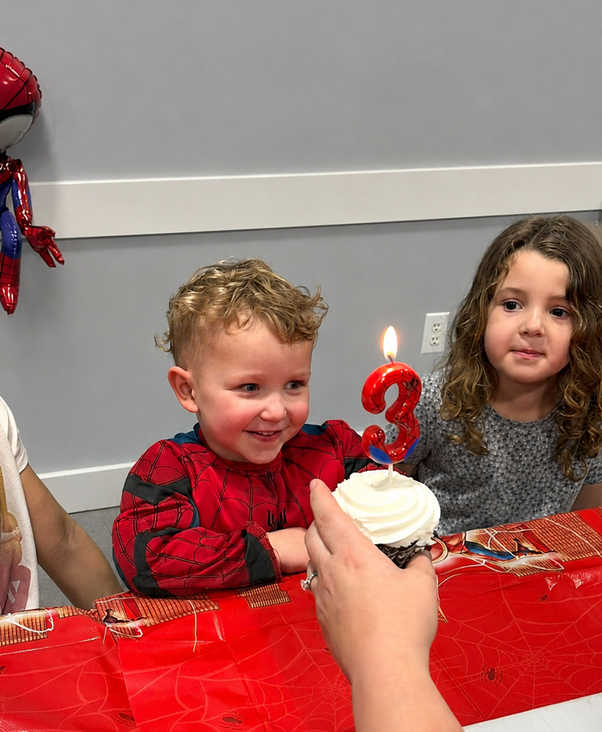 Child at a birthday table with a cupcake and lit number 3 candle, another child watching nearby.