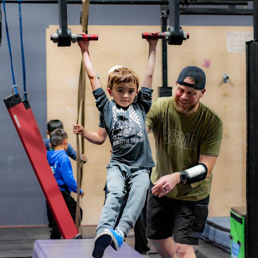 Child on indoor obstacle course swinging from bars with an adult spotting behind him