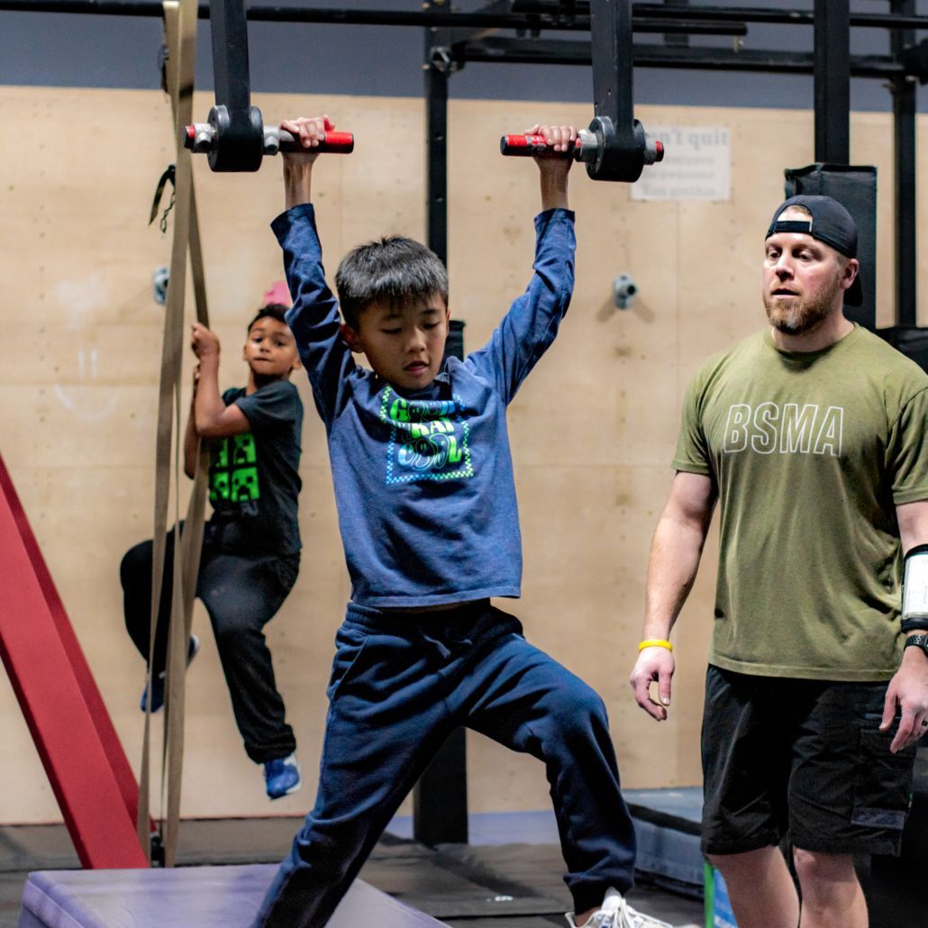 Child hanging from gym rings while a coach stands nearby in an indoor obstacle course.