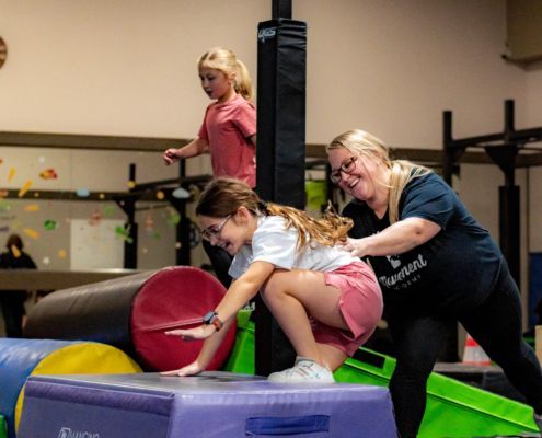 People balancing on foam blocks in a gym, with a coach assisting one child on a mat