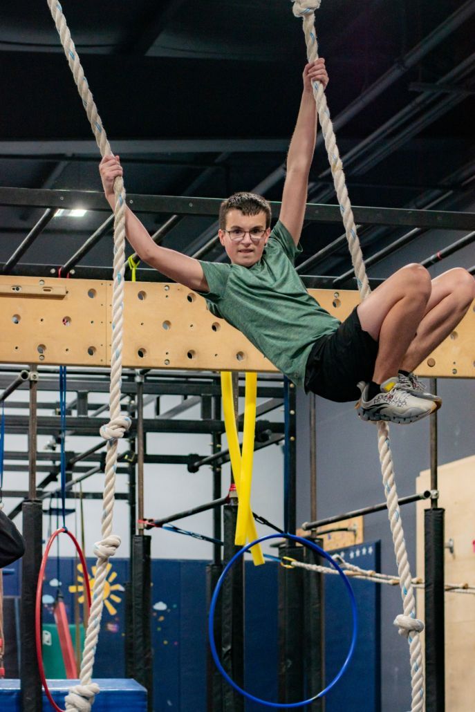 Athlete swings on gym ropes in an indoor obstacle course, wearing a green shirt and black shorts.