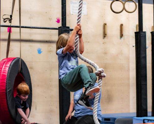 Child climbing a thick rope in an indoor gym while another child watches nearby