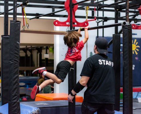 Child in red shirt swings on playground monkey bars while a staff member spots nearby