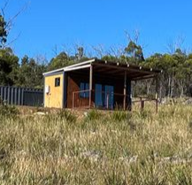 A small cabin with a brown wood exterior and a covered deck, set in a grassy landscape under a clear blue sky.
