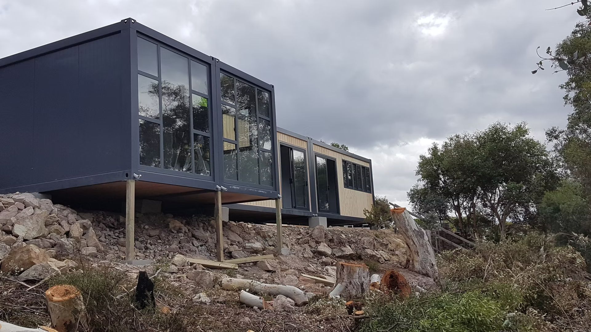A modern, dark-gray modular building with large windows, elevated on stilts on a rocky, uneven hillside under a cloudy sky.