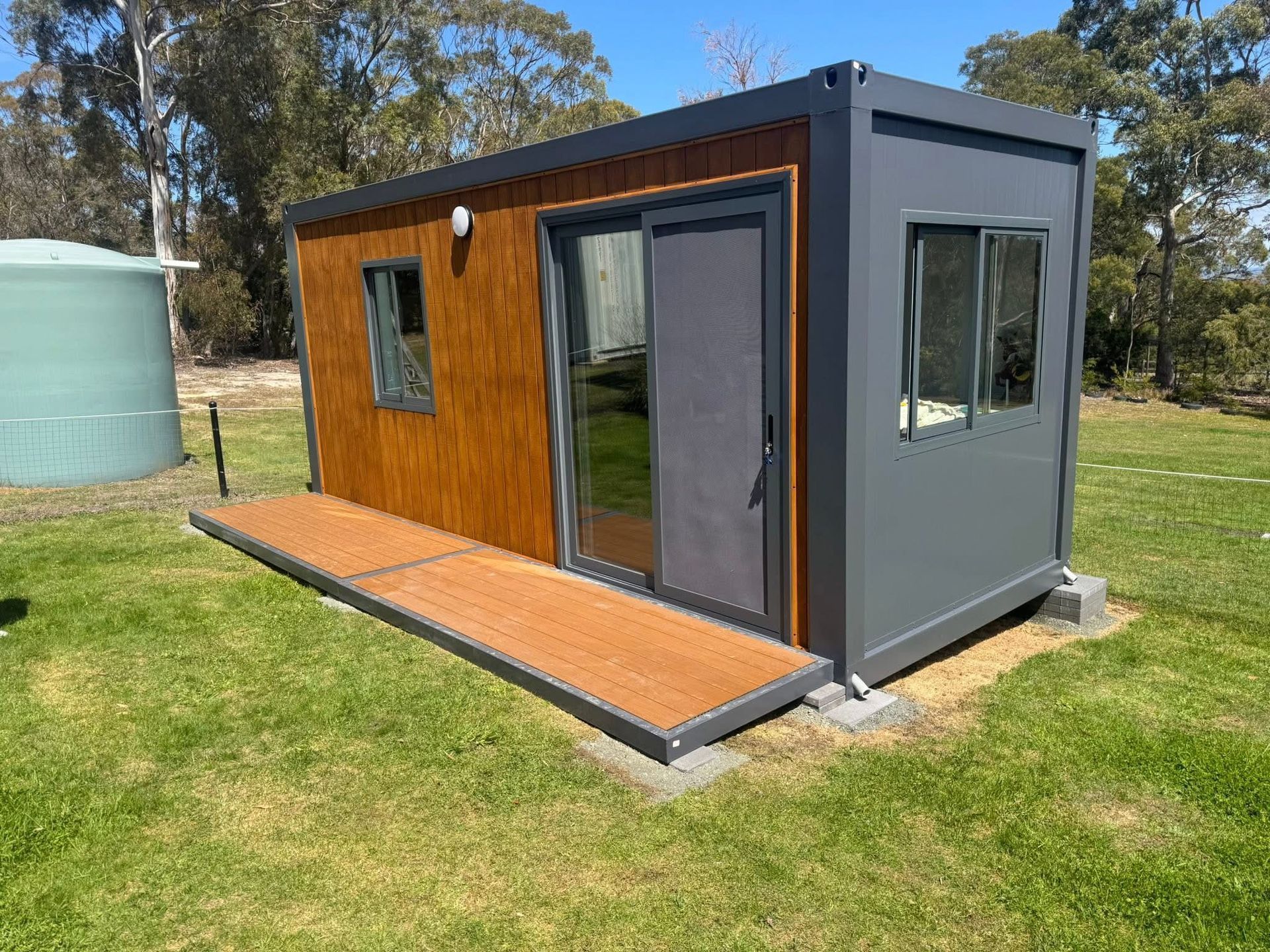 A small, modern wooden cabin with a covered deck sits in a sunny, grassy field surrounded by trees under a clear blue sky.