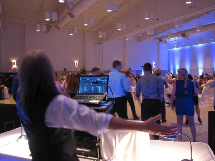 Person at DJ booth performing in a blue-lit event hall with guests gathered on the dance floor