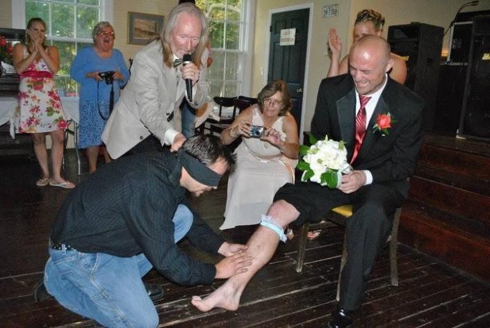 Wedding guests help a seated groom remove a garter during a reception on a wooden dance floor.