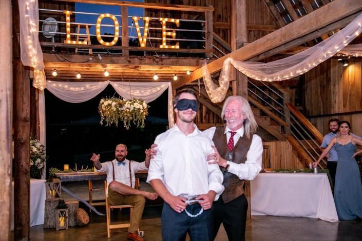 Blindfolded man at rustic wedding reception spins a wheel, with groom-like partner beside him under a LOVE sign