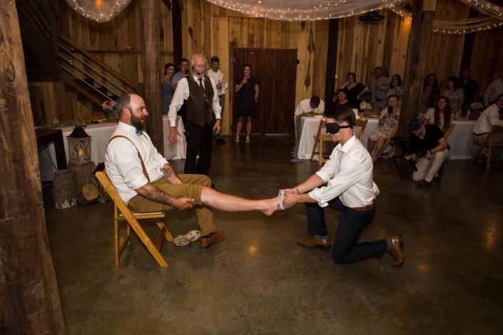 Man kneels and holds another man's foot at a rustic indoor wedding reception with seated guests.
