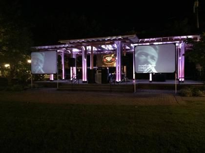 Outdoor nighttime event stage lit in purple, with two large projection screens on a lawn.