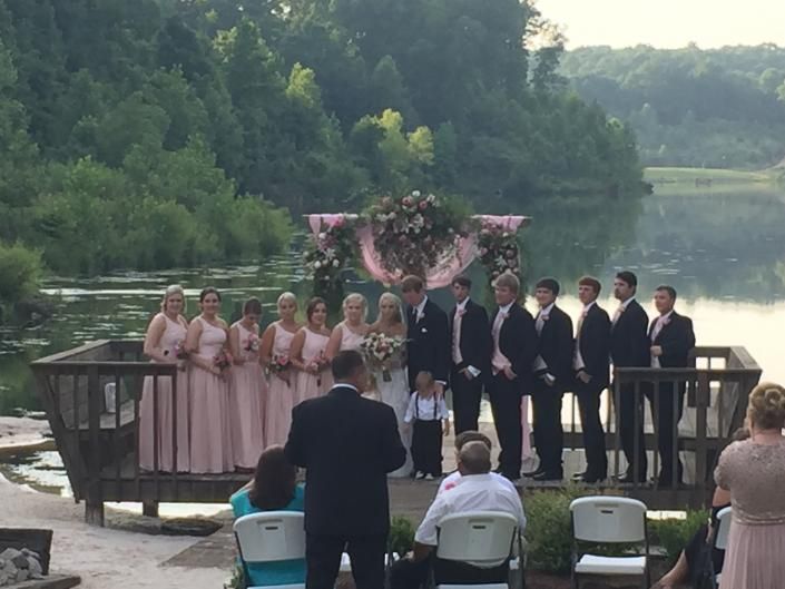 Outdoor wedding ceremony by a lakeside, with bridal party, officiant, and guests under a floral arch.