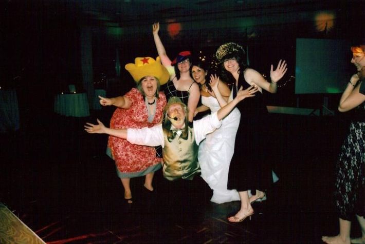 People in costumes dancing at a dimly lit party, with arms raised and colorful outfits.