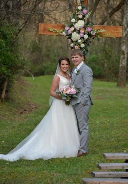 Bride and groom posing outdoors under a wooden sign, holding a bouquet in a grassy wooded setting