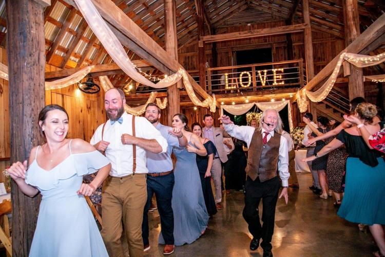 Wedding guests dancing in a rustic barn, with a glowing “LOVE” sign in the background.