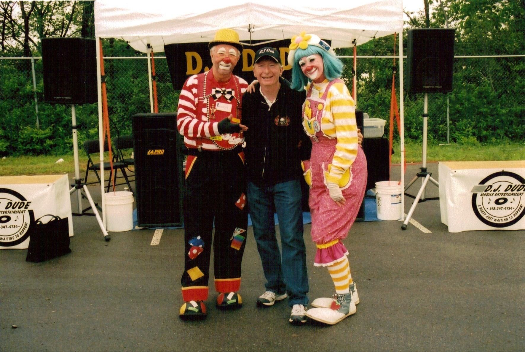 Three people posing on an outdoor stage, two in colorful clown outfits beside a man in black.