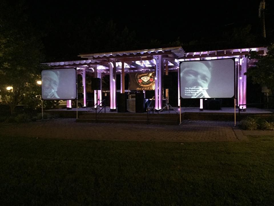 Outdoor nighttime event stage with two large projection screens lit in purple, on a grassy lawn.