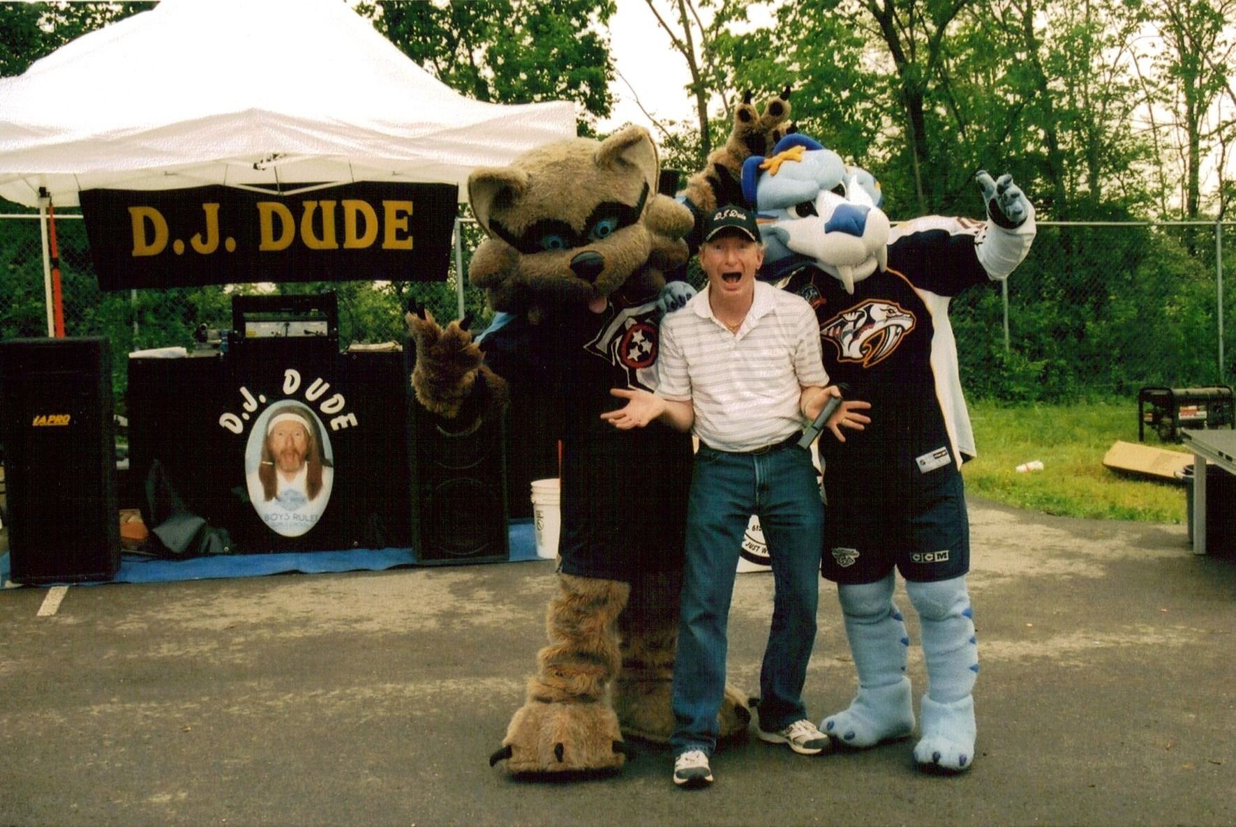 Person posing with two furry mascots beside a D.J. DUDI booth outdoors in a park
