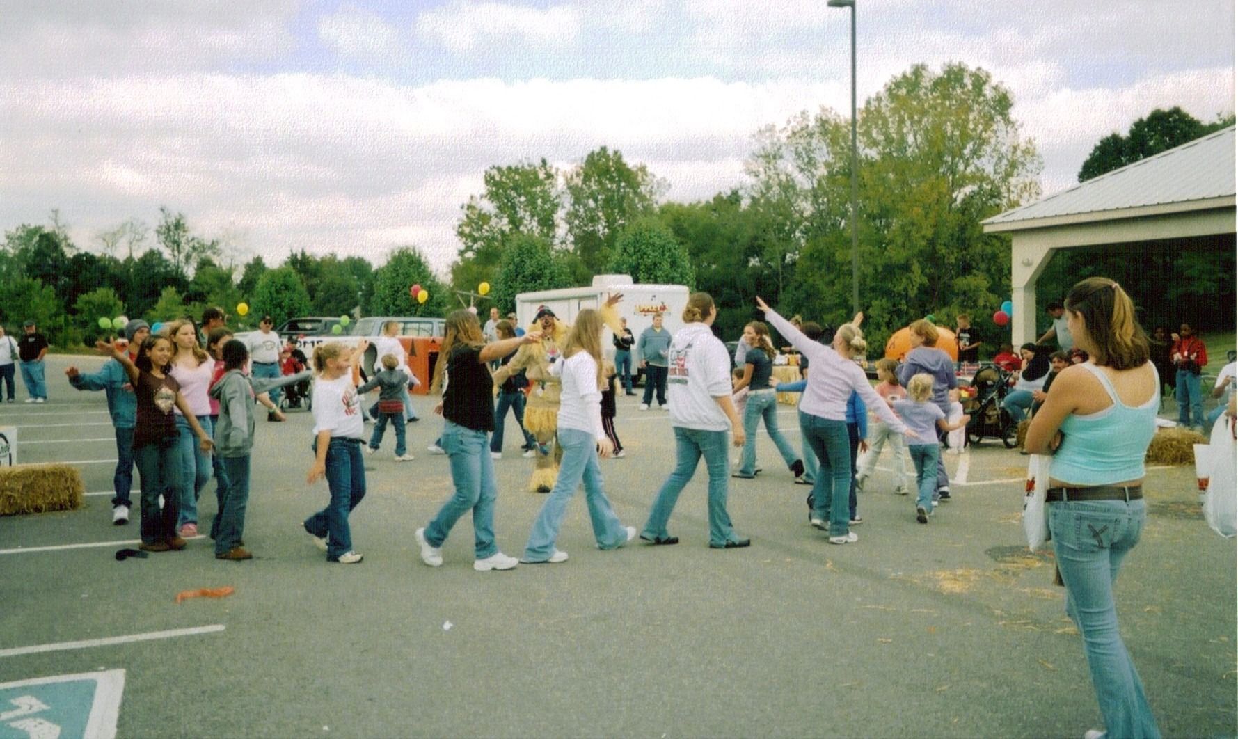 People line dance in a parking lot at an outdoor community event.