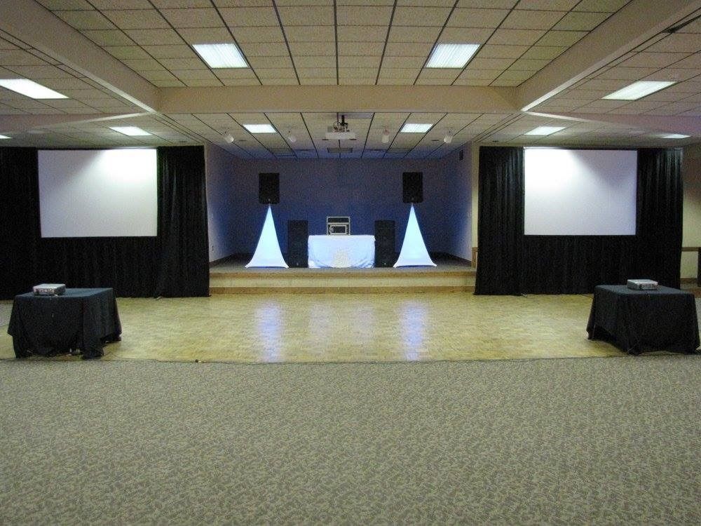 Empty conference hall with stage, two projection screens, and tables under ceiling lights
