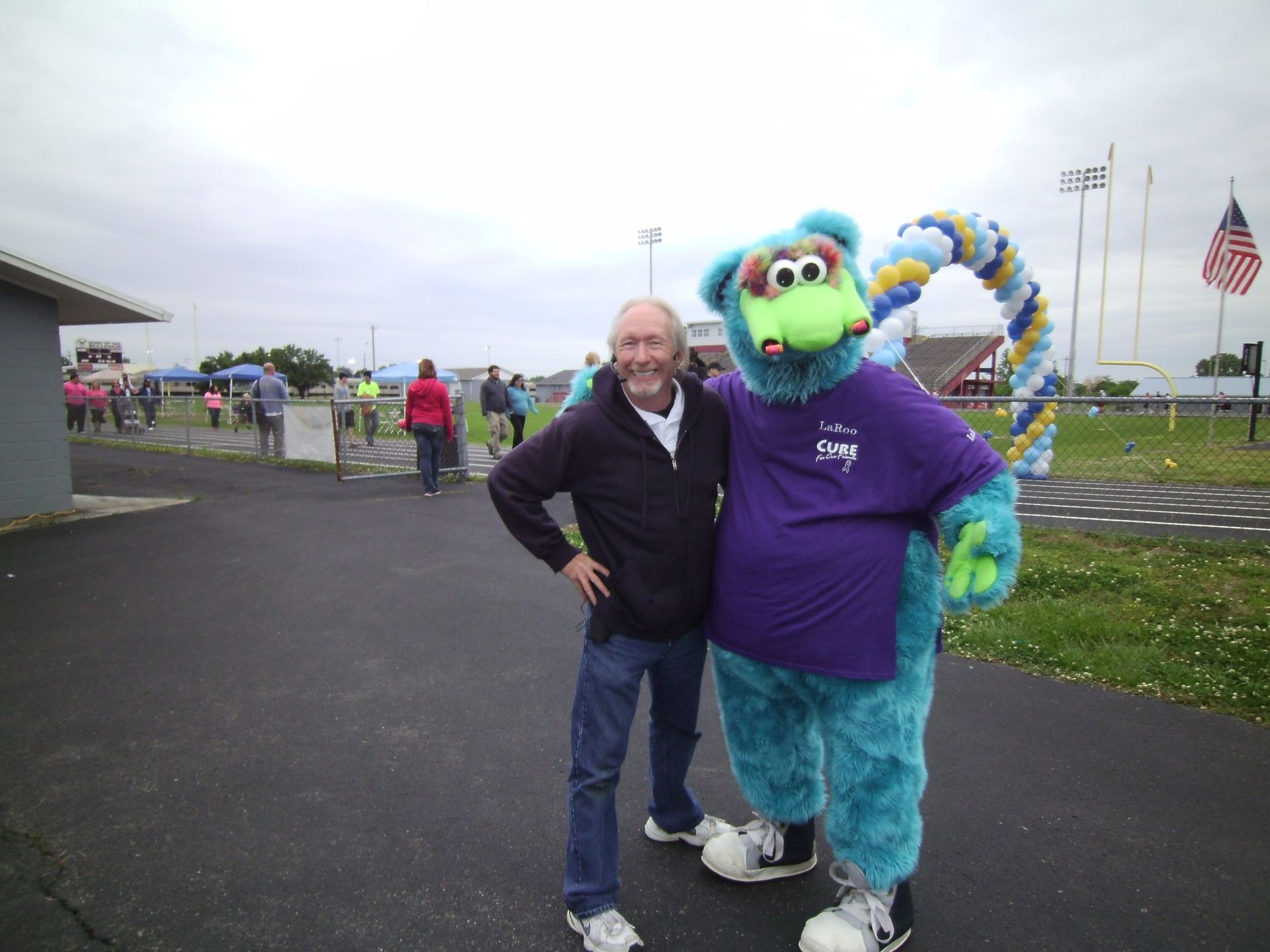 Man posing with a blue mascot in a purple shirt on a paved outdoor lot under cloudy skies